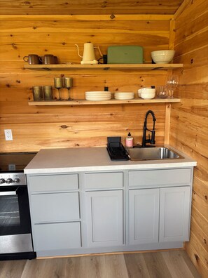 A compact kitchen with wooden walls, a sink, cabinets, and various kitchenware. - The Quiet Acre Cabin (Keswick Ridge)