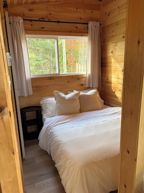 A wooden cabin bedroom with a bed, curtains, and a window overlooking a wooden fence. - The Quiet Acre Cabin (Keswick Ridge)
