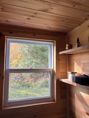 A wooden cabin interior with a window overlooking a forest, a shelf with a bottle, and a container. - The Quiet Acre Cabin (Keswick Ridge)