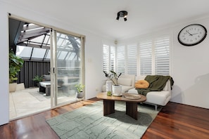 A living room with a sliding glass door leading to a patio, a round coffee table, a sofa, and a wall clock. - New York style meets laid-back Perth living at The Kensington New Yorker.  Enjoy light-filled interiors, beautiful character and a private outdoor space, perfectly positioned near Curtin University,  South Perth, Victoria Park and Perth CBD. (Kensington)