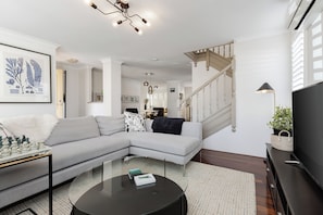 A modern living room with a sectional sofa, a glass coffee table, and a staircase with a wooden railing. - New York style meets laid-back Perth living at The Kensington New Yorker.  Enjoy light-filled interiors, beautiful character and a private outdoor space, perfectly positioned near Curtin University,  South Perth, Victoria Park and Perth CBD. (Kensington)