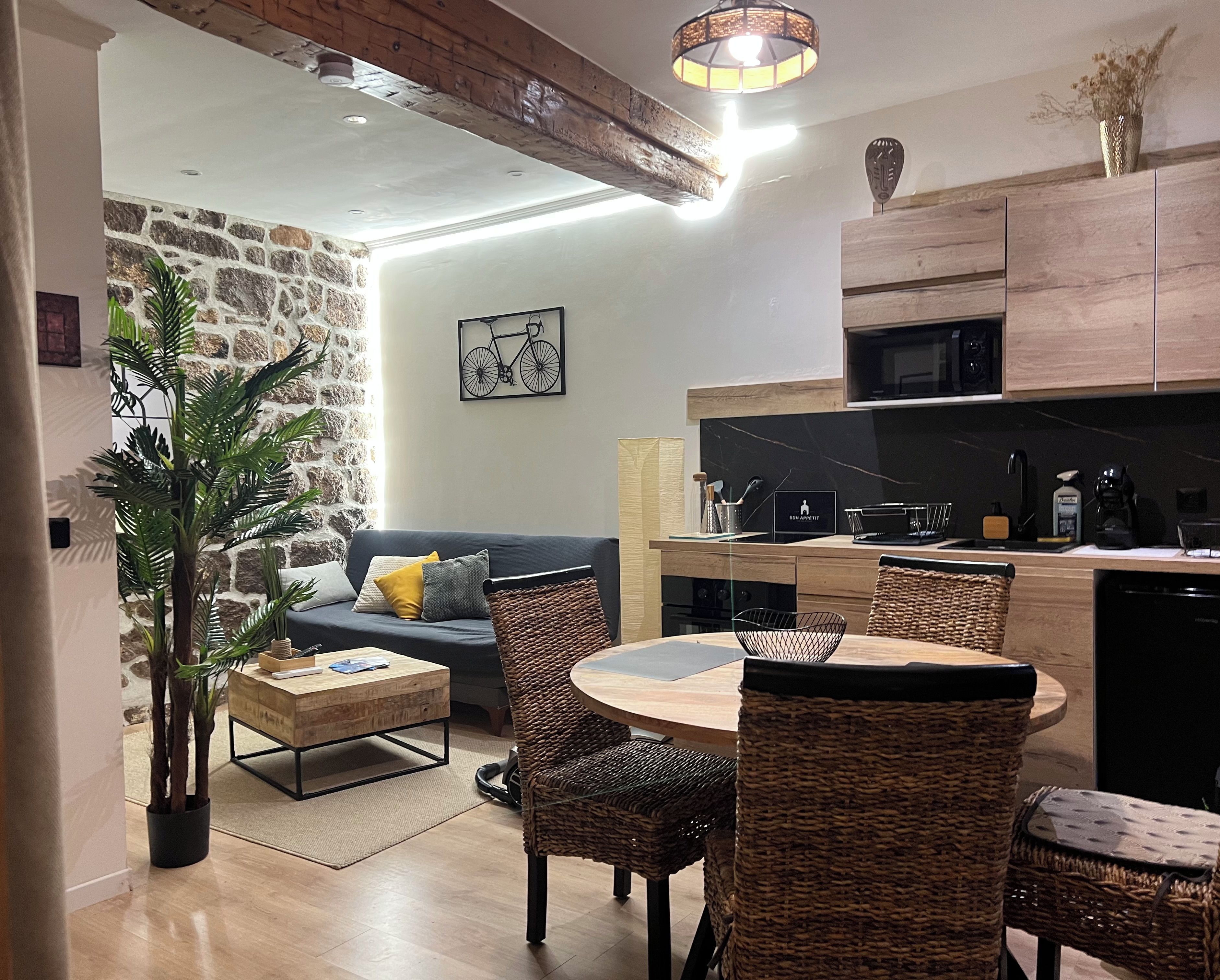 A modern kitchen with a microwave, stove, and wooden cabinets. There is a dining area with a round table and wicker chairs. A potted plant is in the corner, and a wall-mounted light fixture is above the kitchen counter.
