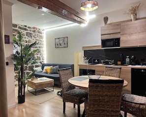 A modern kitchen with a microwave, stove, and wooden cabinets. There is a dining area with a round table and wicker chairs. A potted plant is in the corner, and a wall-mounted light fixture is above the kitchen counter. - An air-conditioned retreat in the heart of Agde's historic center (Agde)
