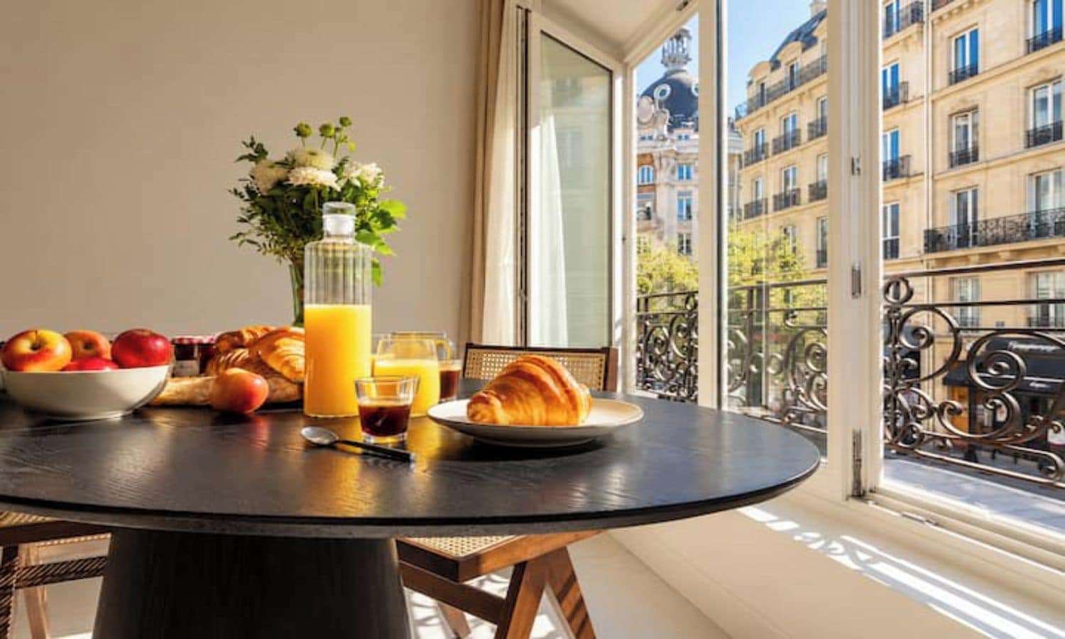 A breakfast setting with a table, fruits, croissants, and a bottle of juice on a balcony with a view of buildings.