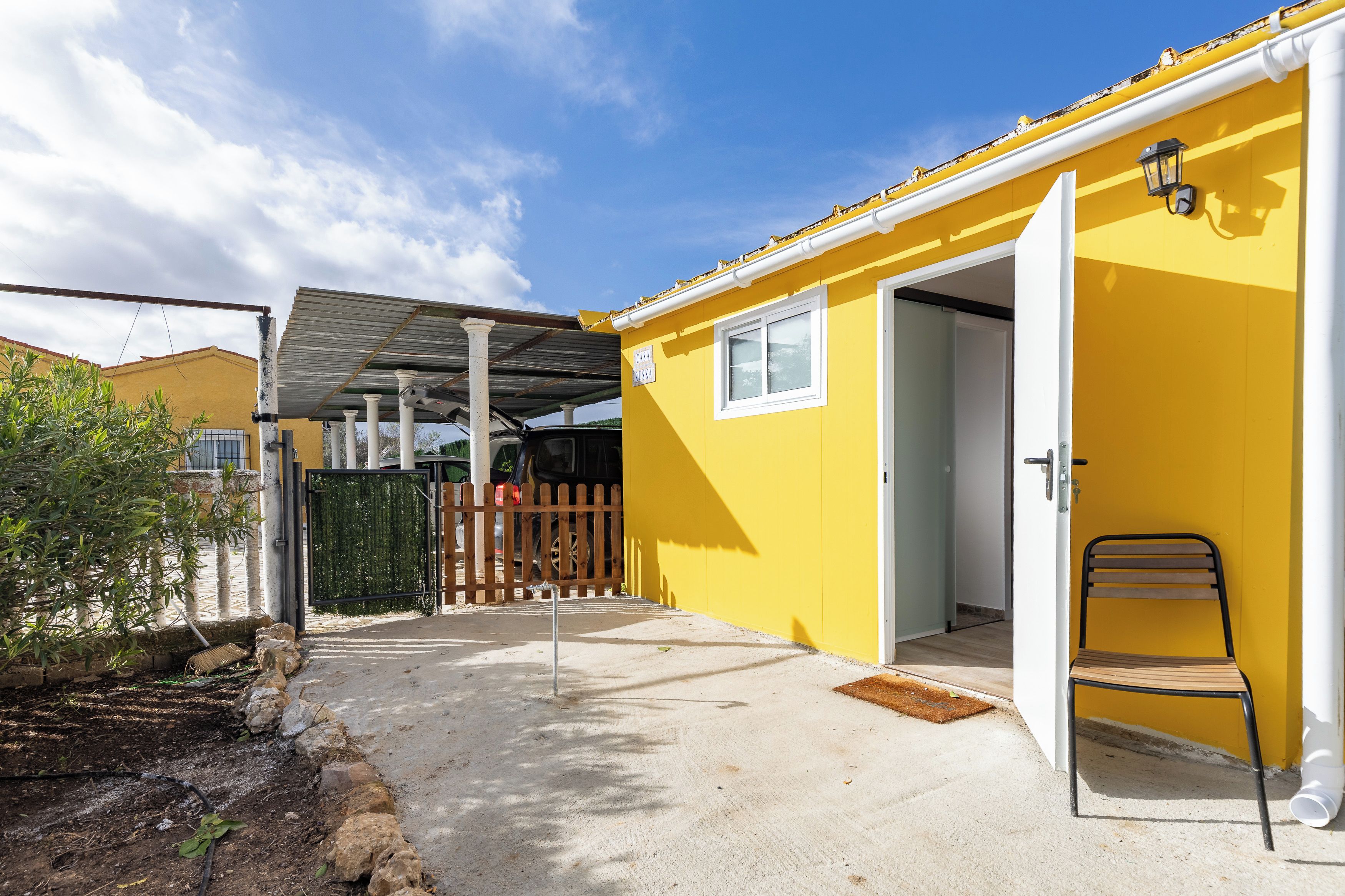 Un edificio amarillo con un patio cubierto, cerca de madera y una silla afuera.