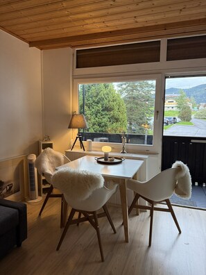 Ein Zimmer mit Holzbalkendecke, einem Tisch mit Stühlen, einer Lampe und Blick auf eine grüne Landschaft durch ein Fenster.