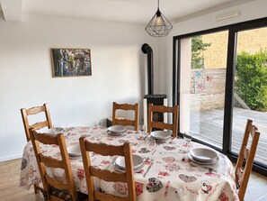 A dining area with a round table, six wooden chairs, and a tablecloth with a pattern. There is a painting on the wall, a black chimney, and a window with a view of greenery. - Across the Fields (Duttlenheim)