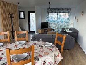 A dining area with a table set for four, a grey sofa, and a television in the background. - Across the Fields (Duttlenheim)