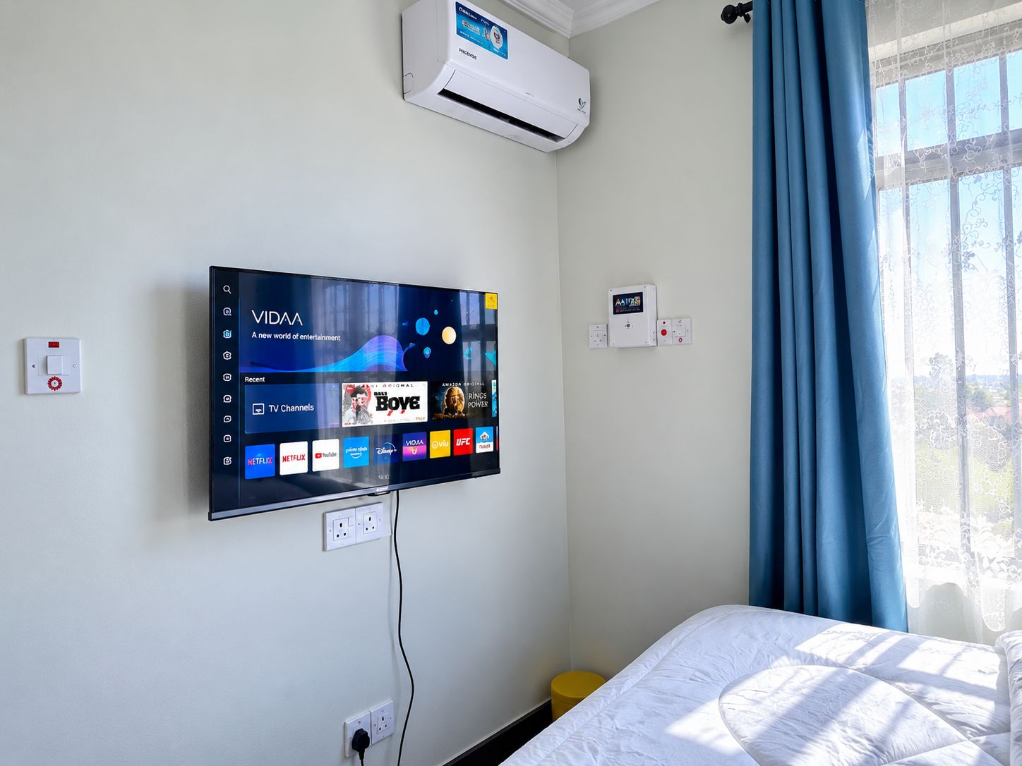 Ein Hotelzimmer mit einem fest installierten Flachbildfernseher, der verschiedene Streaming-Dienste anzeigt, einer Wandklimaanlage und einem Fenster mit blauen Vorhängen.