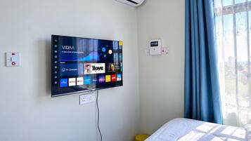 Ein Hotelzimmer mit einem fest installierten Flachbildfernseher, der verschiedene Streaming-Dienste anzeigt, einer Wandklimaanlage und einem Fenster mit blauen Vorhängen.