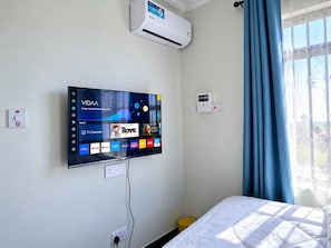 Habitación de hotel con una televisión de pantalla plana montada en la pared que muestra varios servicios de transmisión, un aire acondicionado instalado en la pared y una ventana con cortinas azules.