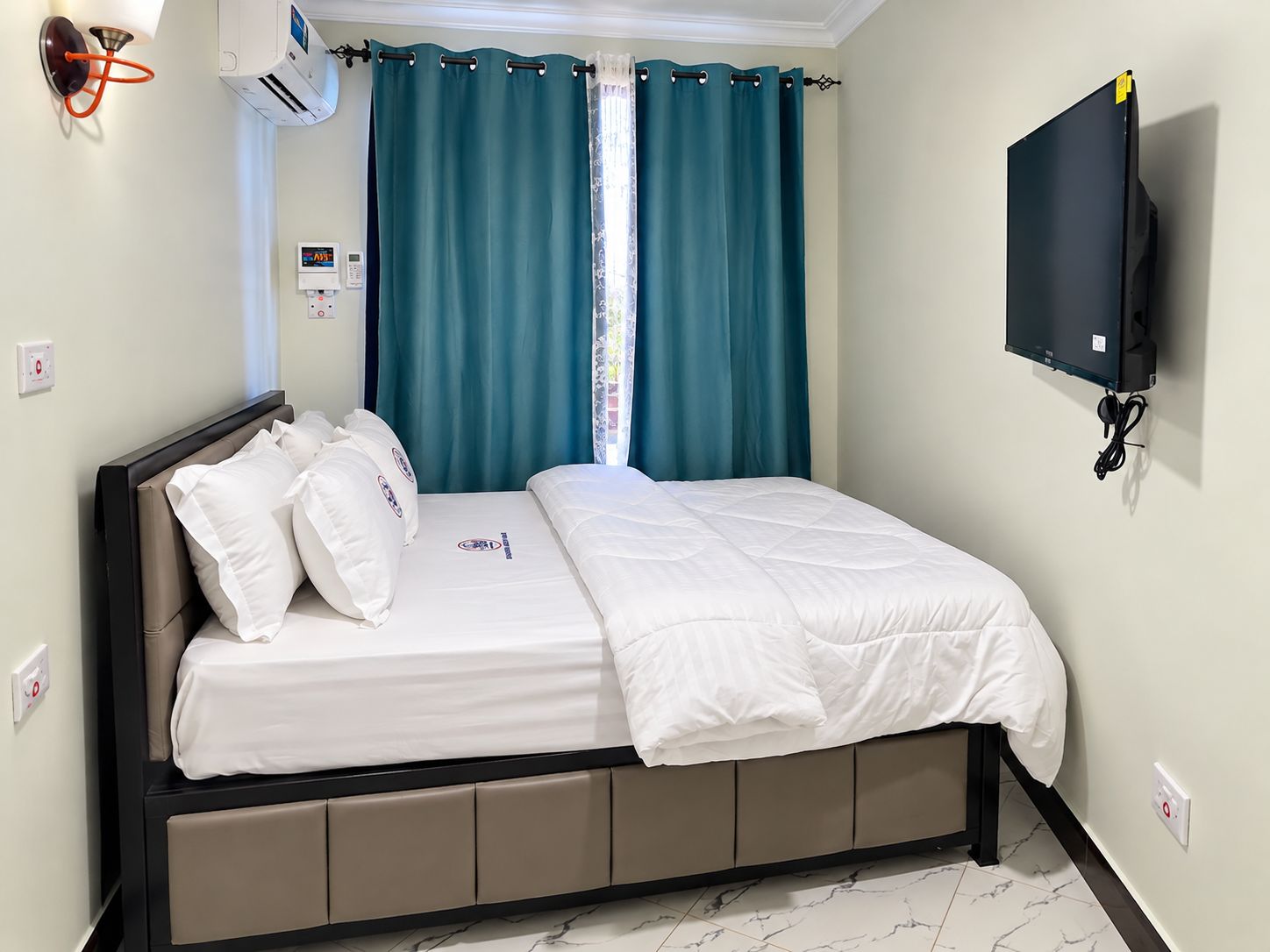 Una habitación de hotel con cama, cortinas, televisor montado en la pared y termostato.
