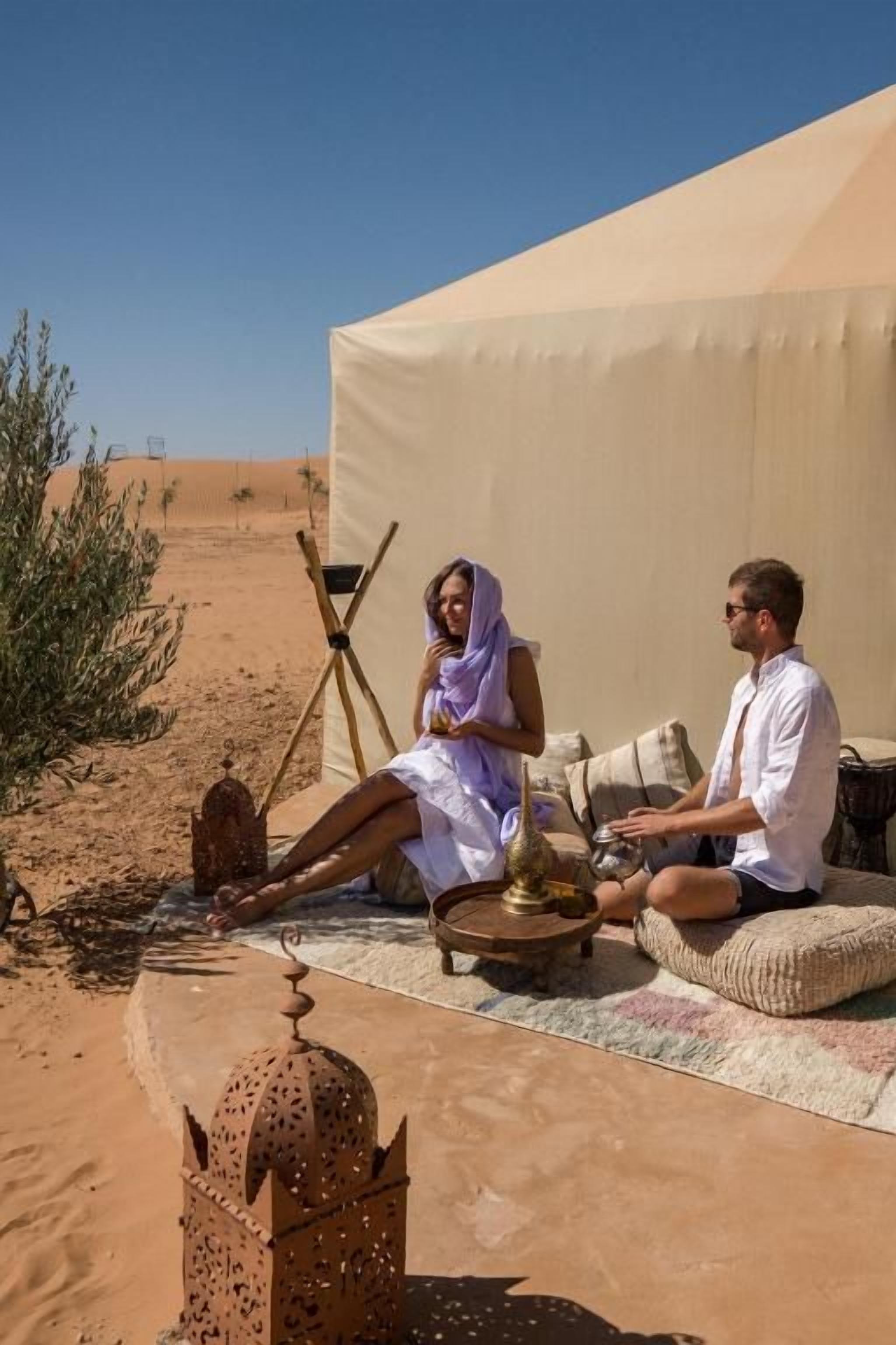 Une tente dans le désert, avec deux personnes assises à l’intérieur, une petite table sur laquelle sont posés quelques objets et une lanterne décorative.