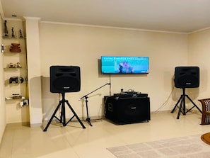 Una sala de estar con un televisor montado en la pared, dos parlantes sobre pedestales, un mezclador de DJ y un estante con objetos decorativos.