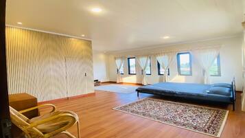 A spacious bedroom with a large bed, a wooden floor, a woven chair, and a view of the outdoors through windows.