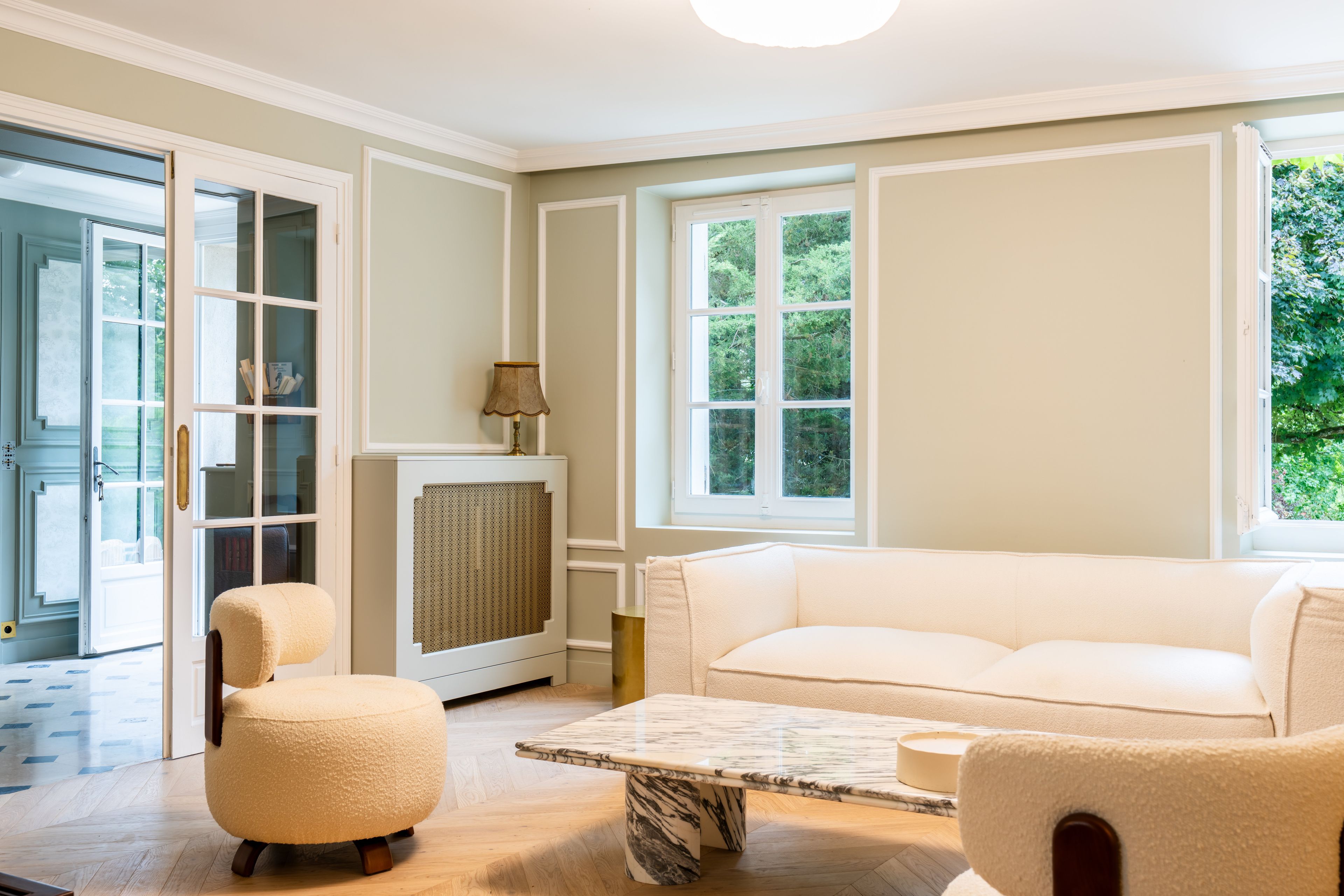A room with a white sofa, a round ottoman, a marble coffee table, and a fireplace with a lamp.