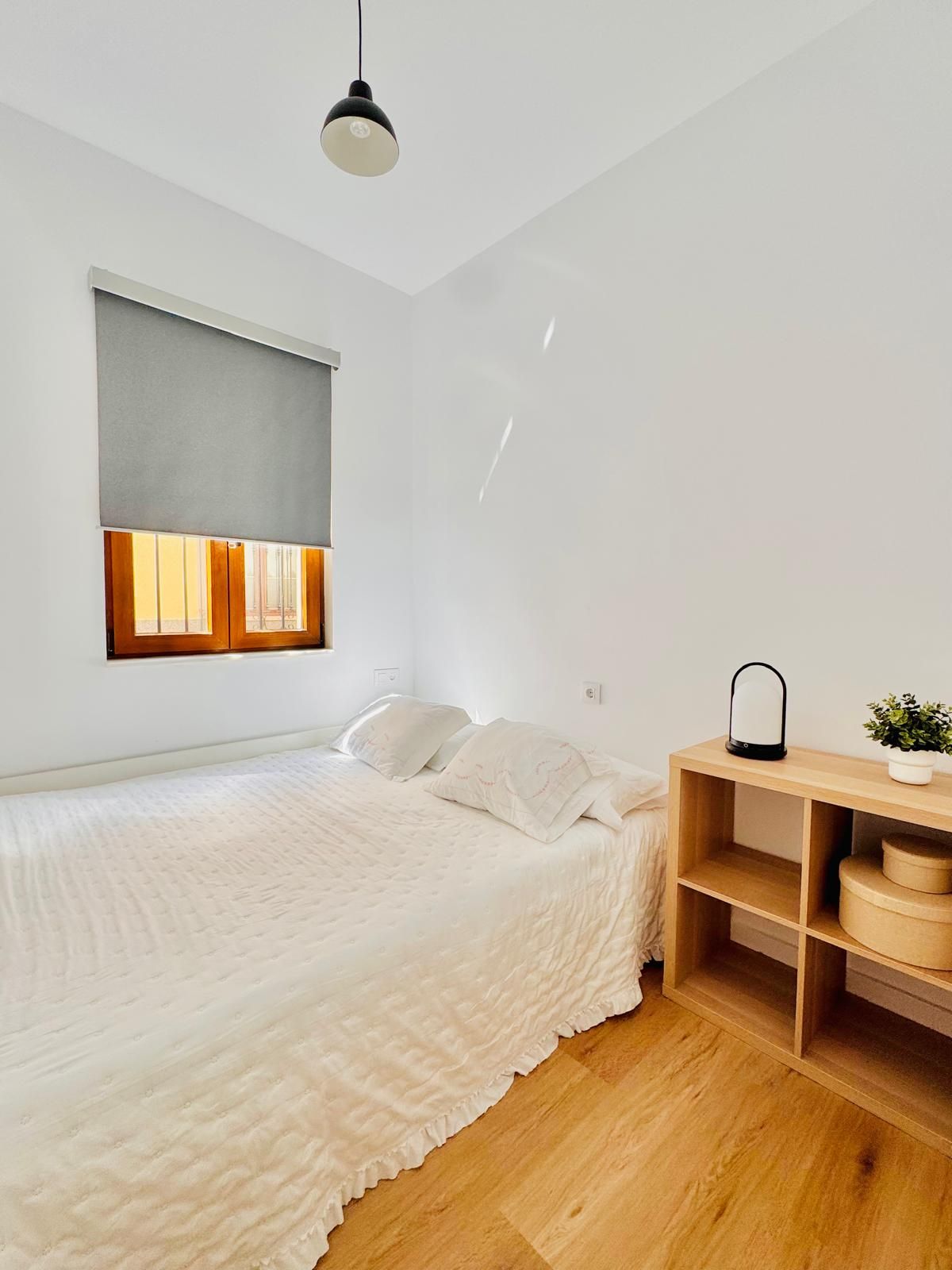Une chambre à coucher comprenant un lit, une table de chevet avec une lampe, une fenêtre avec des stores et une étagère fixée au mur.