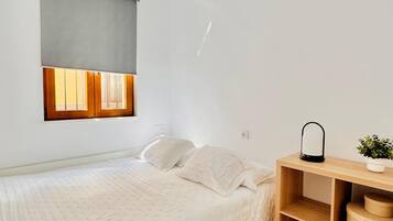 Une chambre à coucher comprenant un lit, une table de chevet avec une lampe, une fenêtre avec des stores et une étagère fixée au mur.