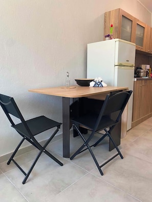 A compact kitchen with a wooden table and two black chairs, a white refrigerator, and a small cabinet with a vase on top. - Brand new House with Veranda for family 2 minutes from the sea (Agia Triada)