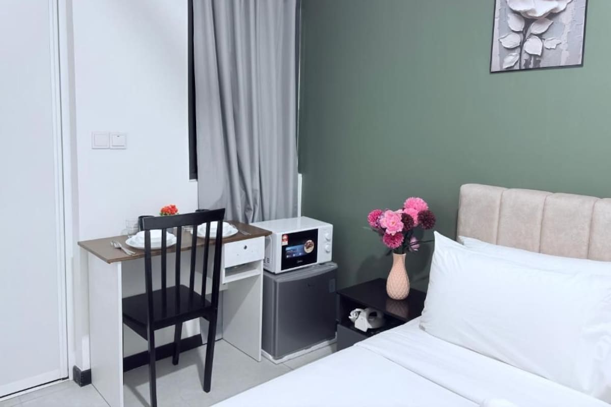 A hotel room with a bed, a desk with a chair, a microwave, a nightstand with a vase of flowers, and a painting on the wall.