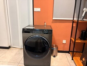 A compact washer and dryer unit in a room with a tiled floor, a small trash bin, and a shelf with a white object. - Neu suite Standard Room By Rasbo Galaxy (Kuala Lumpur)