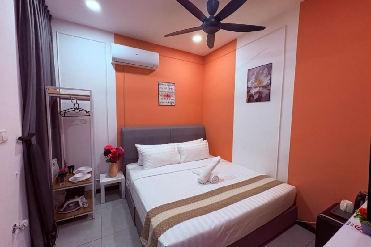 A hotel room with a bed, a ceiling fan, an air conditioning unit, a small table with a vase of flowers, and a wall with a poster.