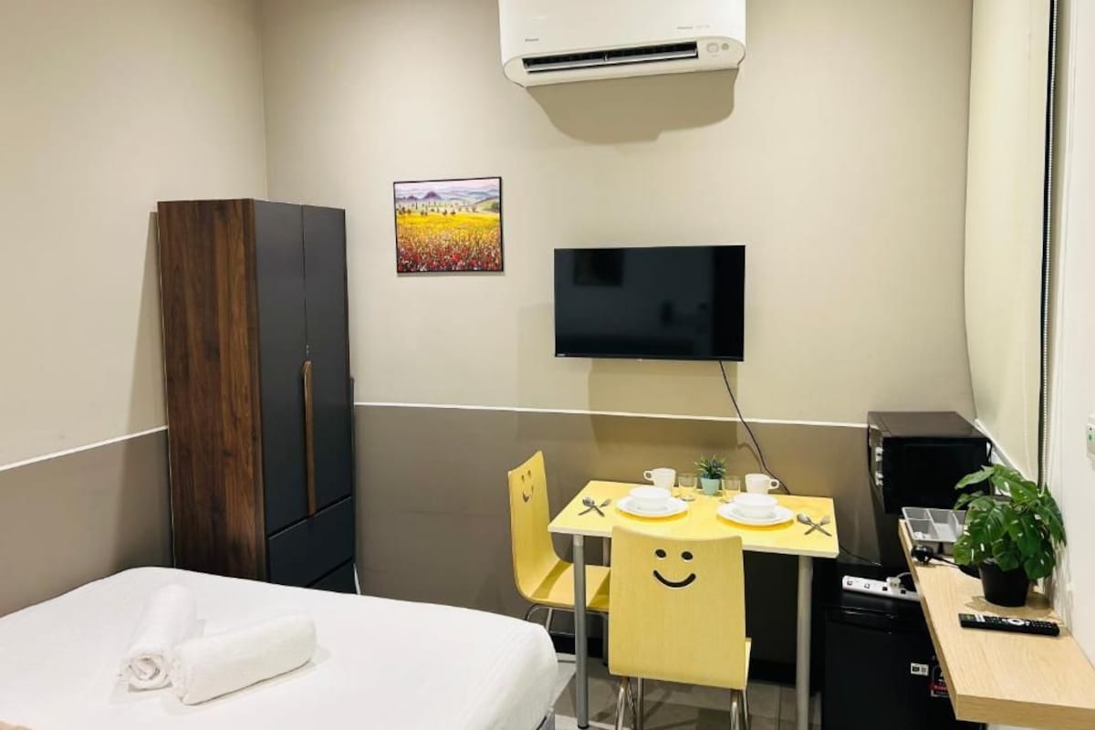 A hotel room with a bed, a desk with a TV, a small table with a plant, and a wardrobe.