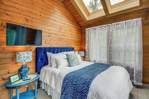 Ein Schlafzimmer mit einem Bett, einem an der Wand befestigten Fernseher, einem Fenster mit durchsichtigen Vorhängen und einem kleinen Tisch mit Lampe.