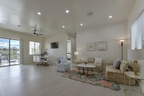 A spacious living room with a dining area, large windows, ceiling fan, and a flat-screen TV mounted on the wall.
