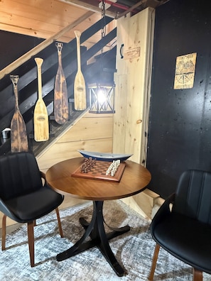 Una habitación con una mesa de madera, dos sillas, un tablero de ajedrez y una maqueta de barco.