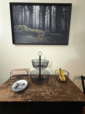 A dining table with a framed forest landscape, a two-tiered wire basket, a plate with a photo, and a banana. - Serene 5-bedroom house in delightful Old Forge perfect for families (Old Forge)