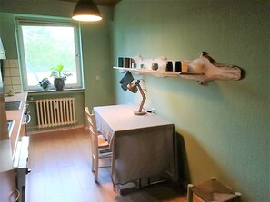 A kitchen with a table, chairs, a hanging light, a shelf with kitchen items, and a window with plants. - Vacation Rental with an Extra-Large Natural Garden & Sunny Terrace (Clausthal-Zellerfeld)