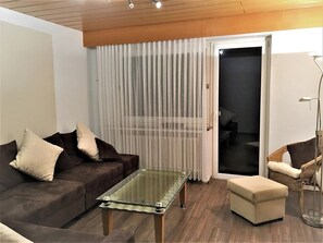 A living room with a glass coffee table, a brown sofa with cushions, a wicker chair, and a sliding glass door leading to a balcony. - Vacation Rental with an Extra-Large Natural Garden & Sunny Terrace (Clausthal-Zellerfeld)