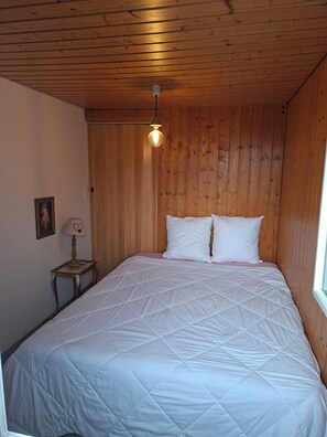Un dormitorio con techo de madera, una cama con ropa blanca, una mesita de noche con lámpara y un cuadro enmarcado en la pared.