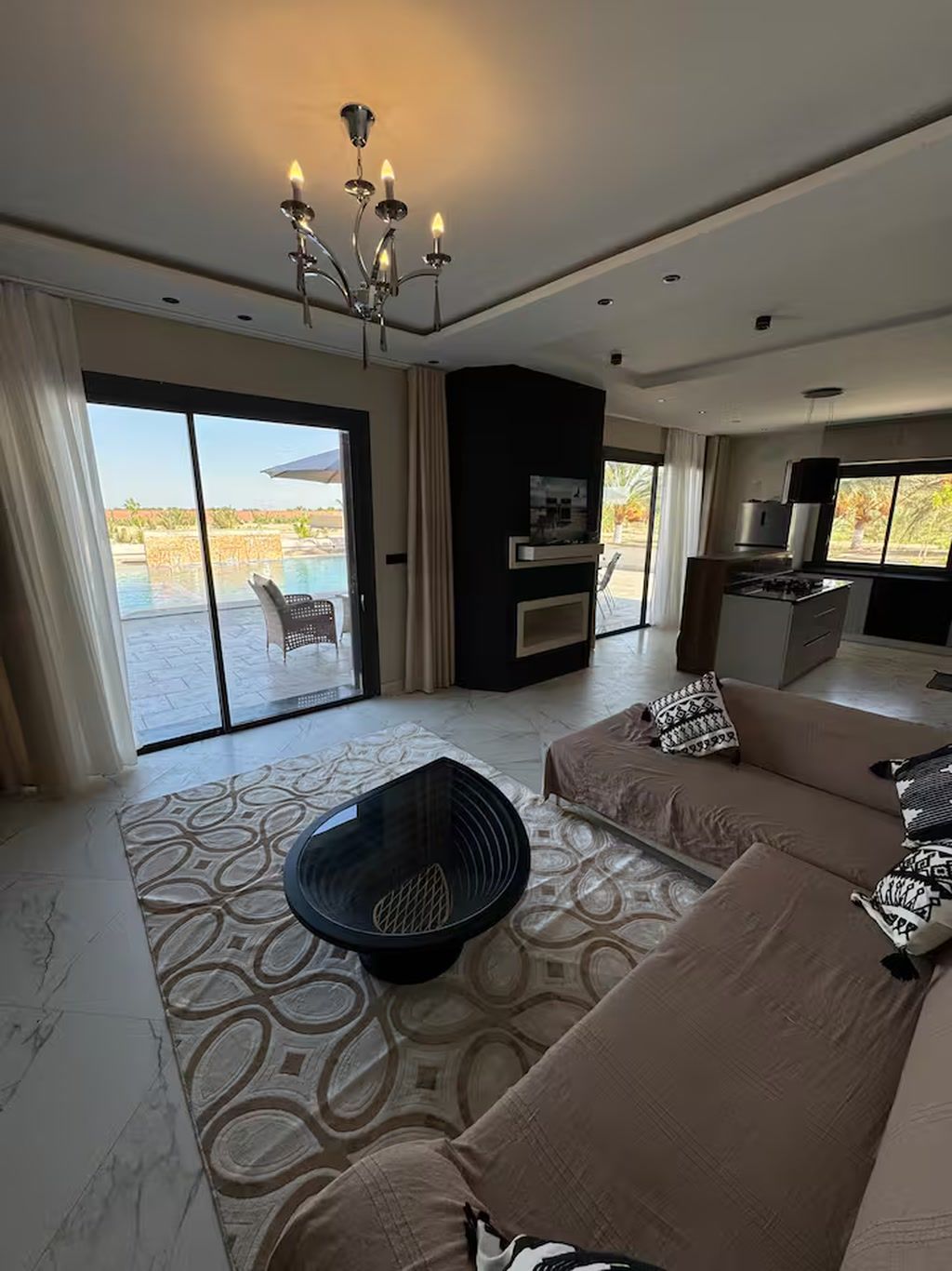Una sala de estar moderna con un amplio sofá, una mesa de centro negra y una chimenea. Hay una puerta corrediza de vidrio que da a una zona de piscina.
