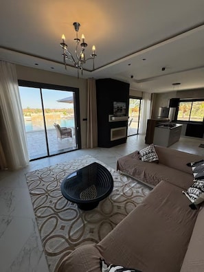 Una sala de estar moderna con un amplio sofá, una mesa de centro negra y una chimenea. Hay una puerta corrediza de vidrio que da a una zona de piscina.