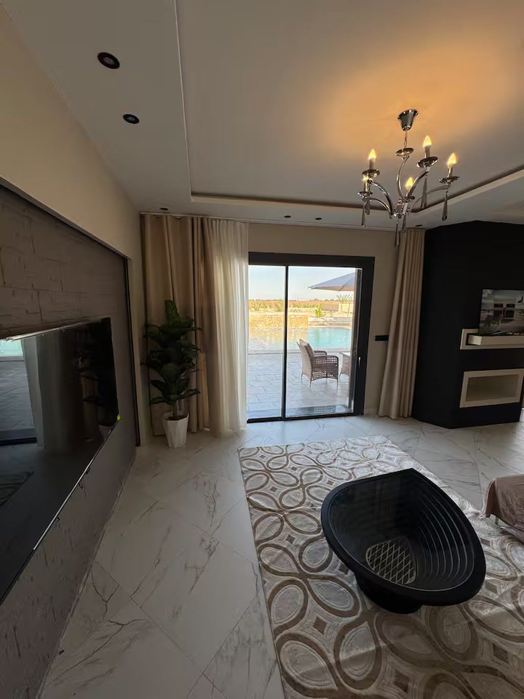 A modern living room with a flat-screen TV, a patterned rug, a black ottoman, and a view of an outdoor pool through a glass door.