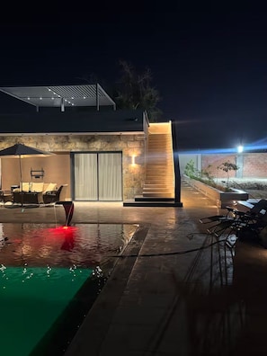 A modern outdoor pool area with a stone wall, a seating area with chairs, and a staircase leading to a covered deck. - Villa in the heart of a palm grove (Mekhadma)