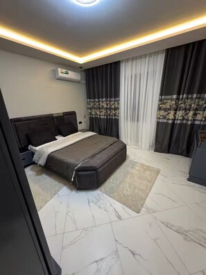 Un dormitorio moderno con una cama grande, cabecera, mesita de noche, aire acondicionado instalado en la pared y una ventana con cortinas.