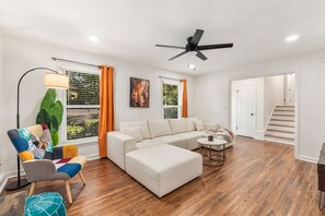 Un salon moderne avec un canapé d’angle, une petite table ronde, un fauteuil coloré et un ventilateur de plafond.