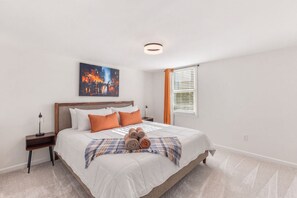 A bedroom with a bed, a nightstand, a lamp, a painting, and a window with blinds. - Movie Theatre Southpark Patio WFH Setup (Charlotte)