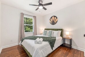 Une chambre à coucher comprenant un lit, un ventilateur de plafond, une fenêtre avec des rideaux, un tableau décoratif au mur et une table de chevet avec une lampe.