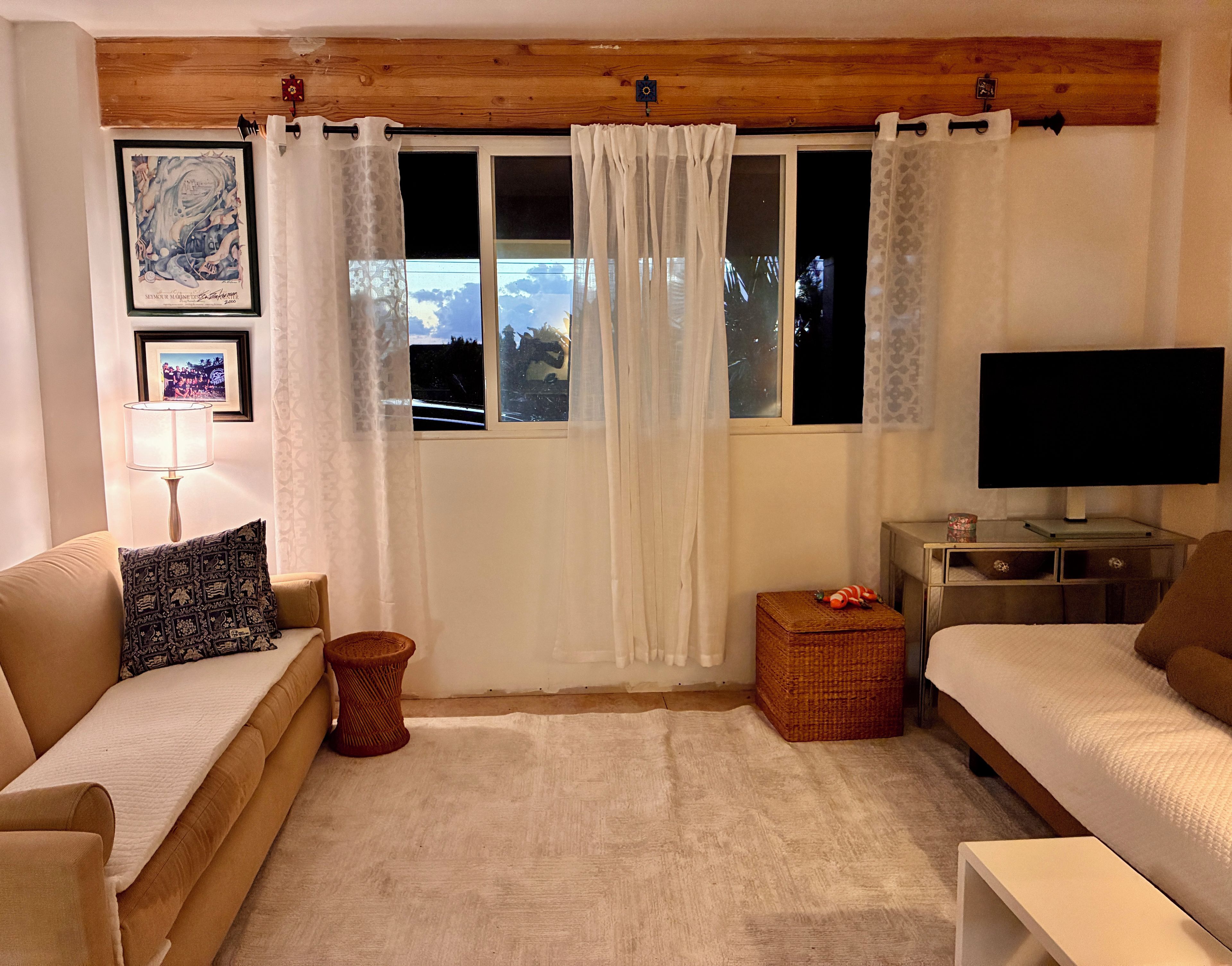A room with a sofa, a television, a window with curtains, a small table, and a wall-mounted picture.