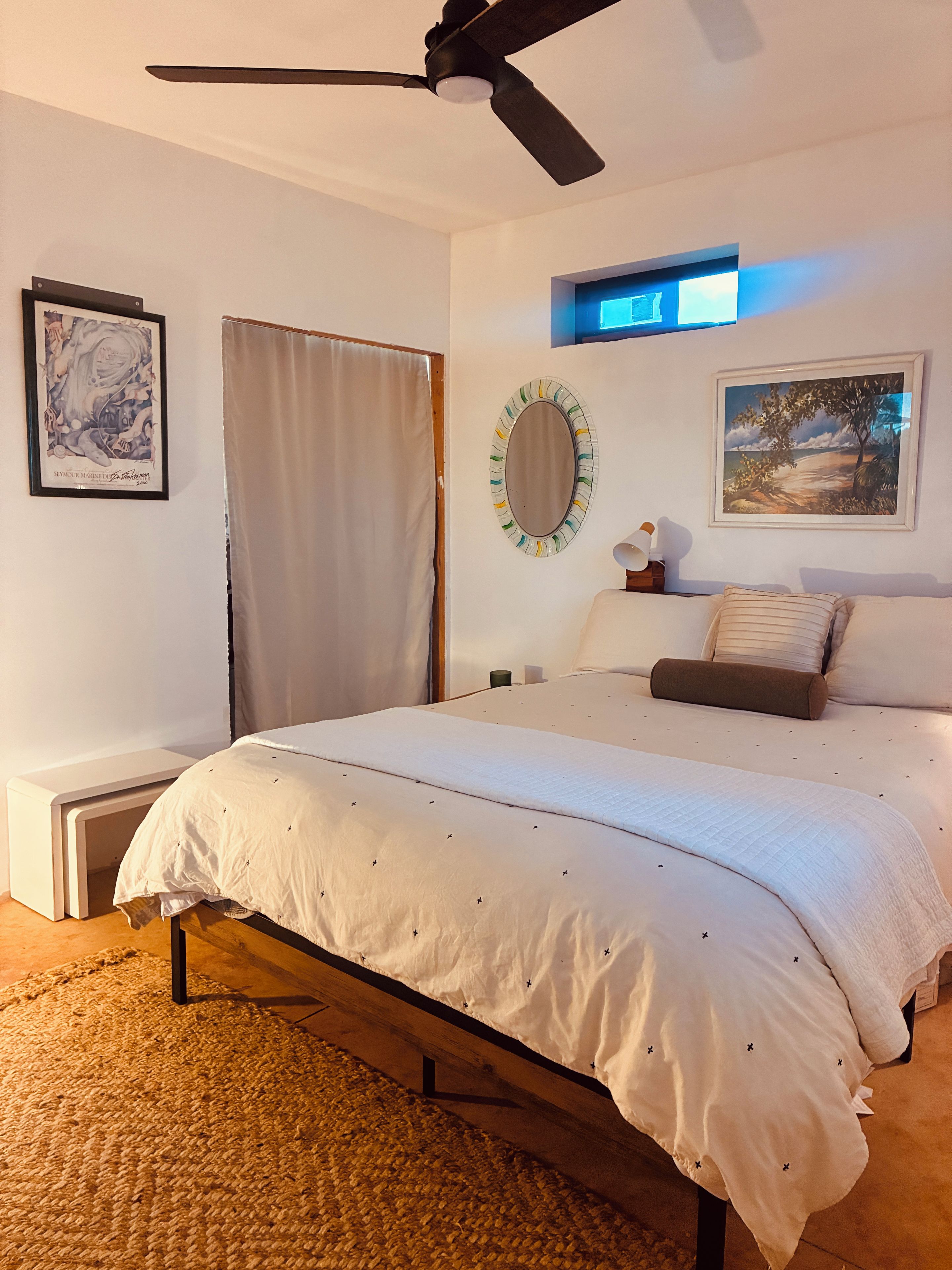 A bedroom with a bed, a ceiling fan, a wall-mounted mirror, a framed picture, and a nightstand.
