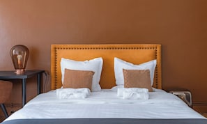 A neatly made bed with white linens and two brown pillows, a bedside table with a lamp, and a brown wall.