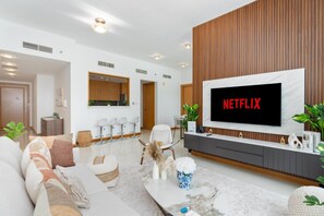 A modern living room with a large flat-screen TV mounted on a wooden wall, a sofa set, a coffee table, and a dining area with chairs. - Warm 2BR Marina View at Iris Blue (Dubai)
