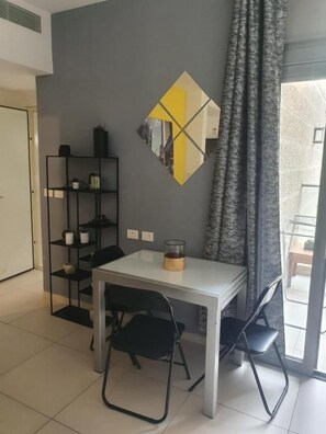 A room with a dining table, chairs, a shelf with items, and a window with curtains. - HaRav Kuk Royal Suites (Jerusalem)