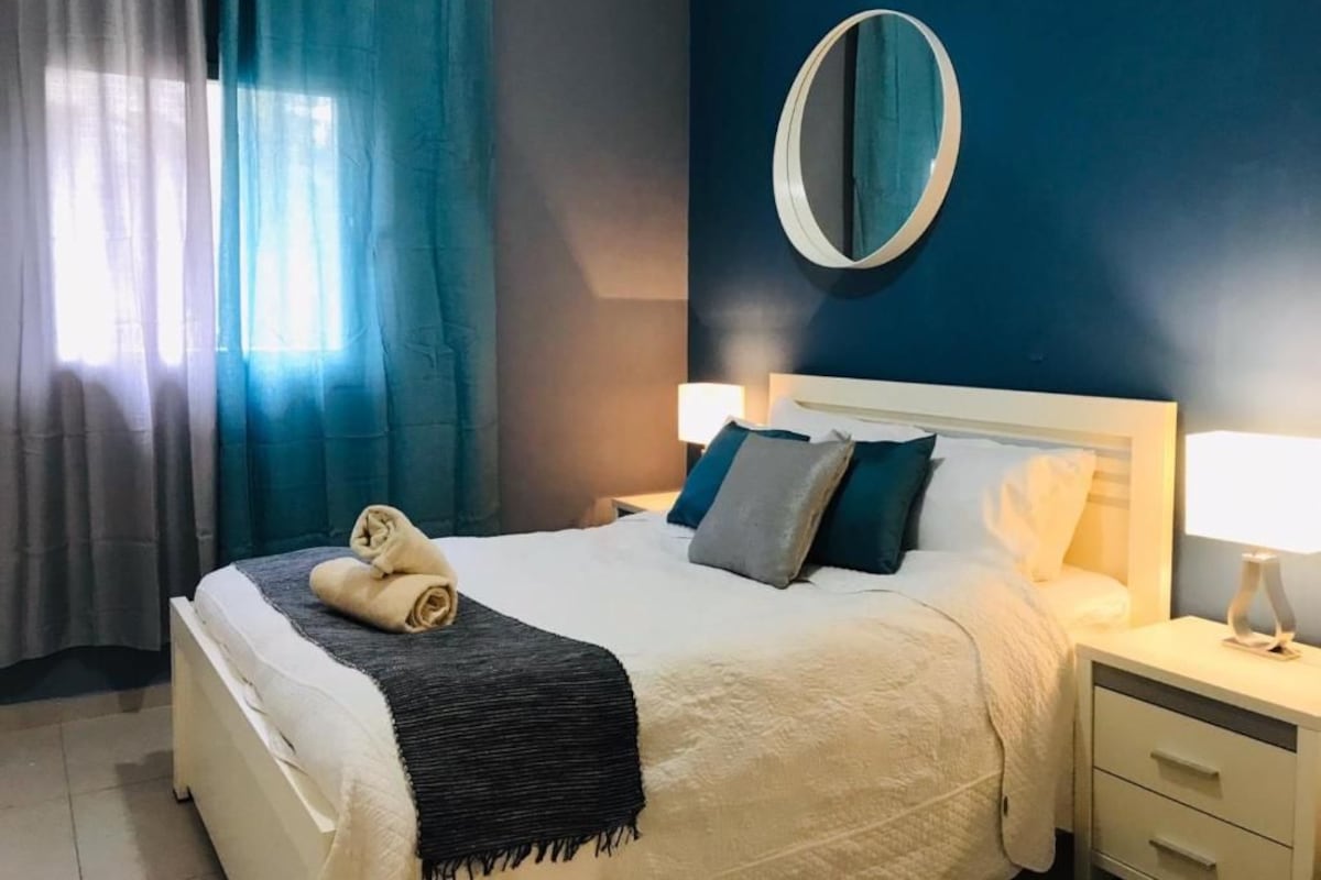 A hotel room with a bed, a bedside table, a lamp, a mirror, and curtains.