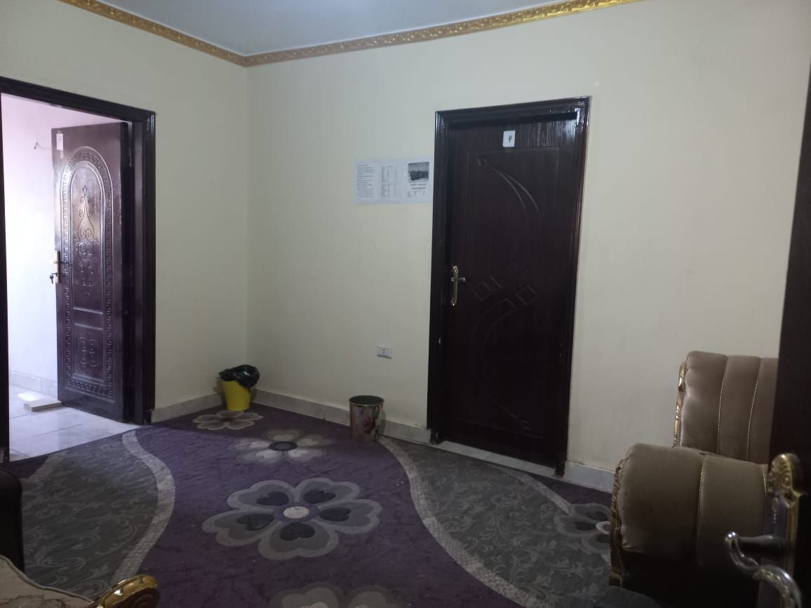 A room with a patterned carpet, a closed door, a trash bin, and a chair.
