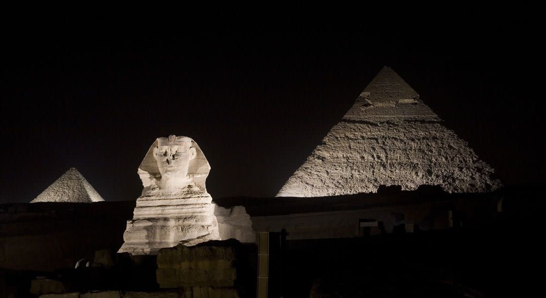 Una estructura piramidal amb una gran cabeça de pedra al primer pla i dues piràmides més petites al fons, il·luminades.